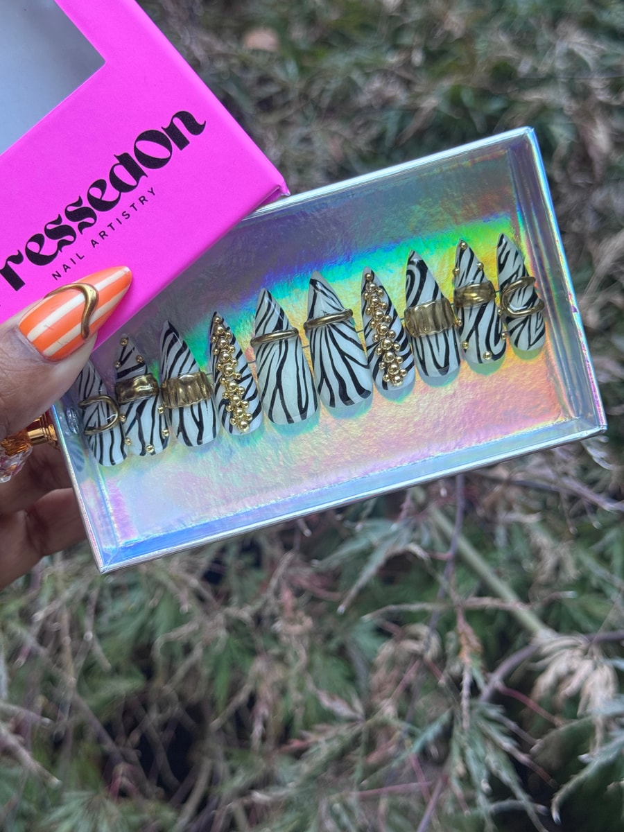 The image showcases medium length press-on nails with a zebra print design. The nails are decorated with gold rings and studded accents.